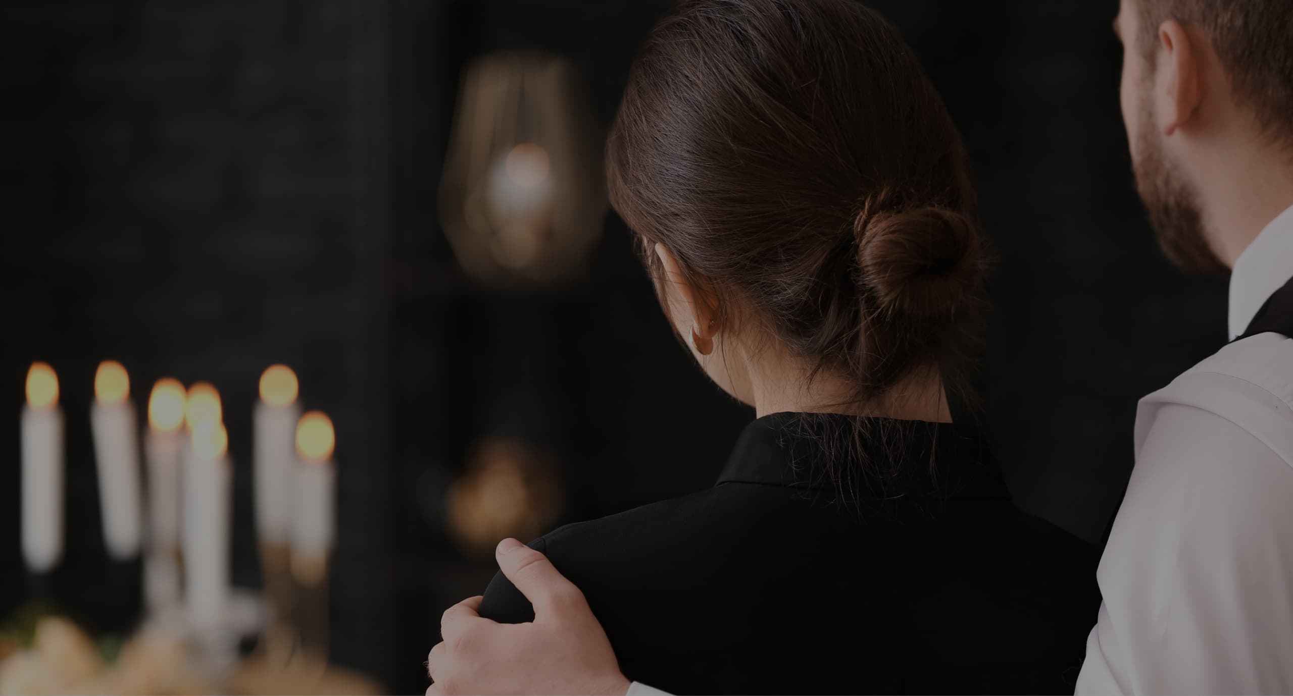 A woman in black standing with a man with his arm around her shoulder staring at candles. Used for multiple pages including, obituaries,